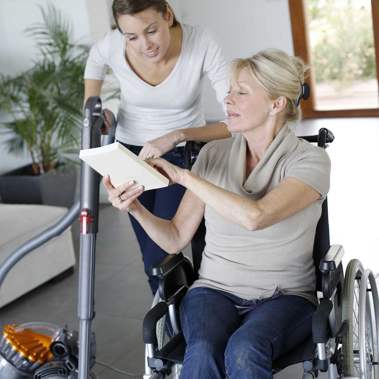 Photo of a caregiver stopping the vacuuming to look at a book to which a senior client is pointing.
